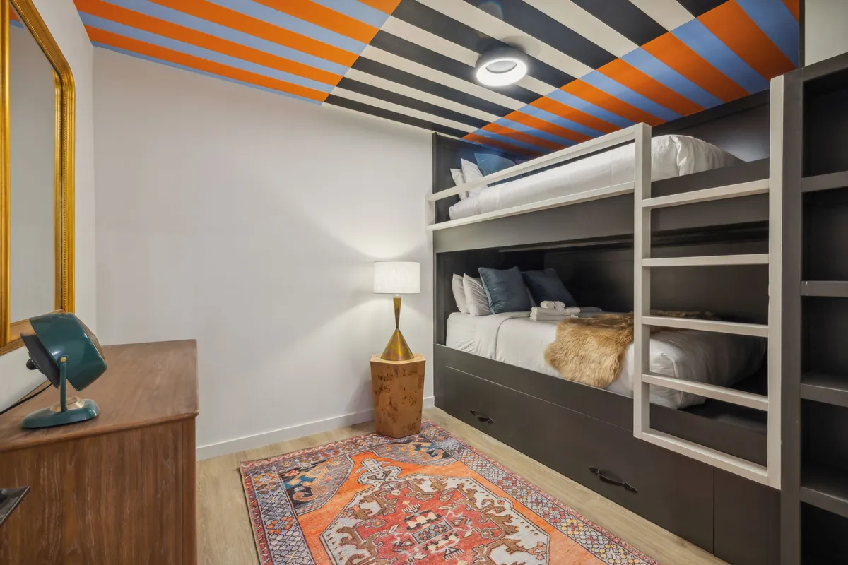 A vibrant bunk room with colorful ceiling details, a bunk bed setup, area rug, lamp, and a dresser. The playful and inviting decor suggests a perfect space for families or groups, offering both fun and comfort.
