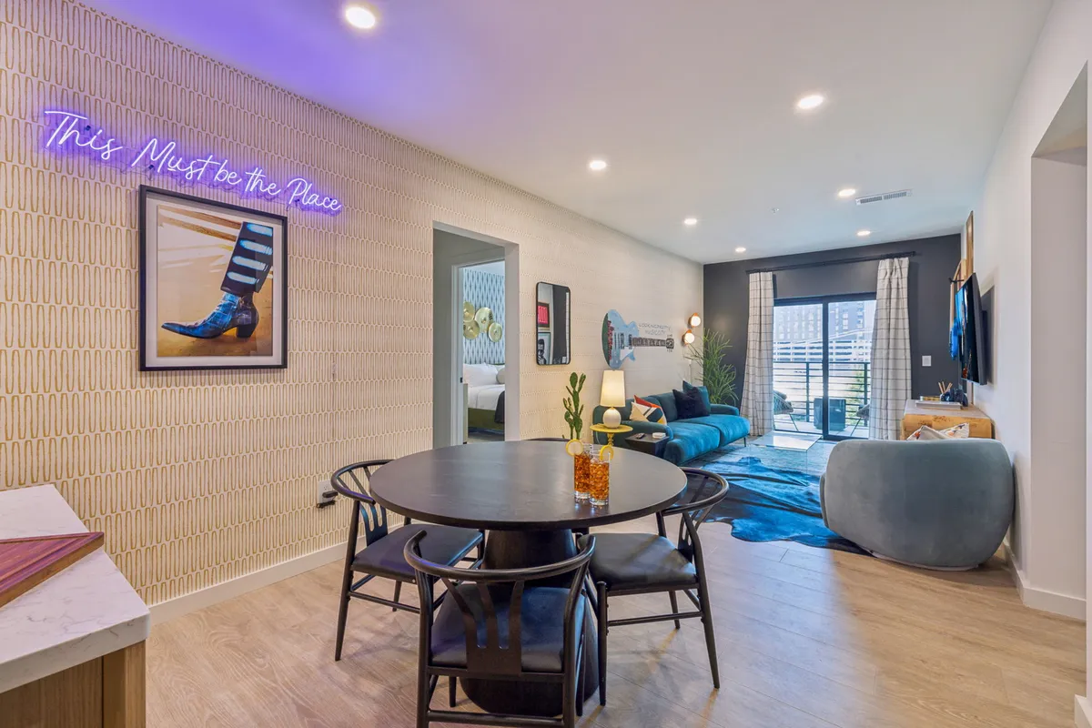 A playful dining area with a neon sign, a spacious table, modern blue couch, and eclectic wall art. The open design and colorful decor foster an atmosphere for shared meals, lively conversation, and entertaining guests.