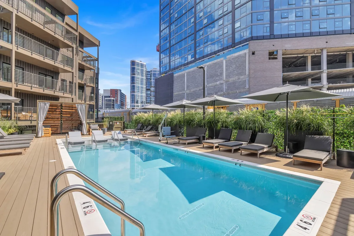 A panoramic view of a swimming pool area featuring plenty of lounge chairs, shade umbrellas, and lush greenery, with high-rise buildings visible in the background. The setting suggests a peaceful yet lively communal space where guests can unwind, socialize, enjoy refreshments, or take in urban views.