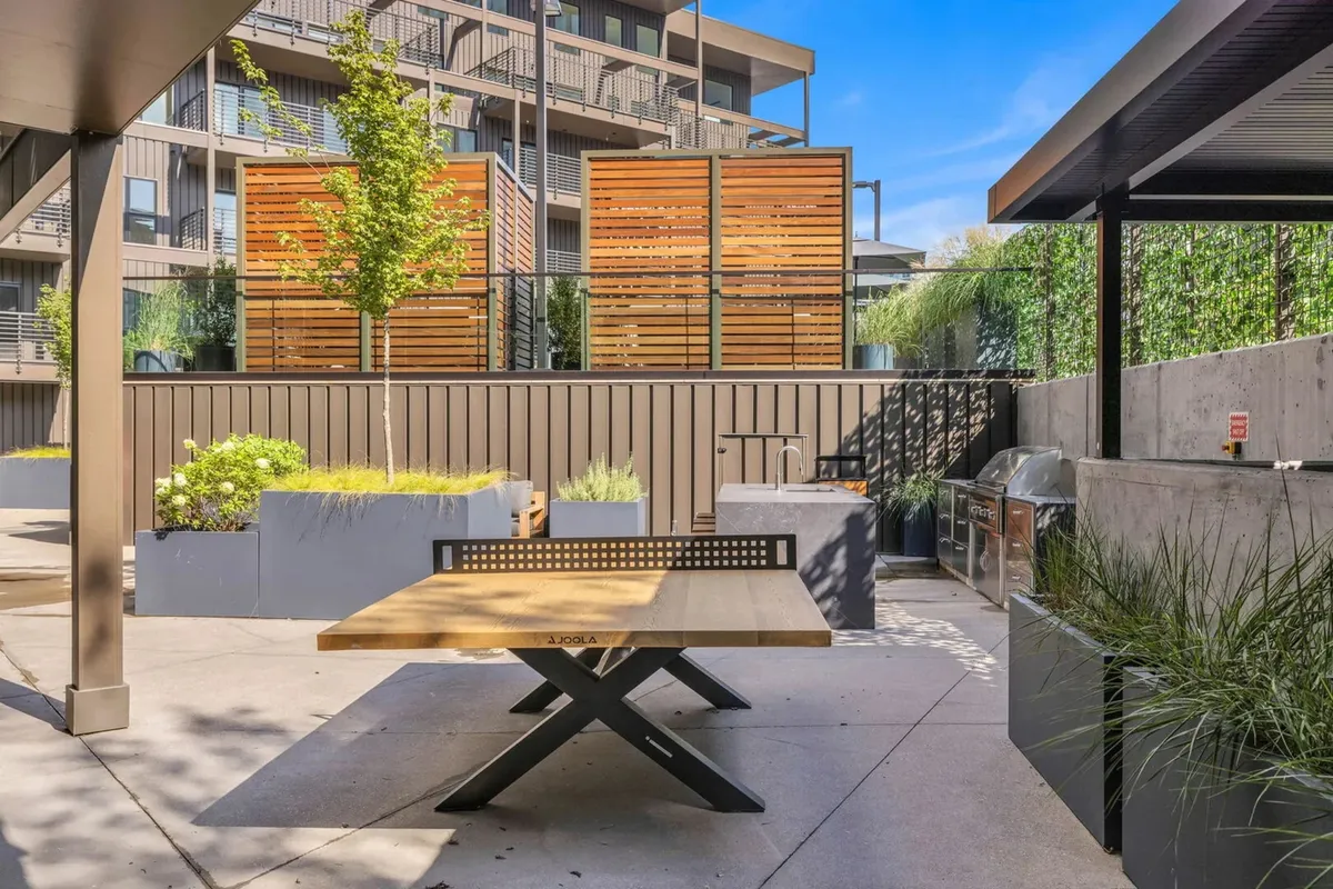 A welcoming outdoor grill area with a wooden dining table, decorative screens for privacy, and green plants. The space is designed for al fresco dining, barbecuing, and social gatherings, evoking a casual and communal vibe.