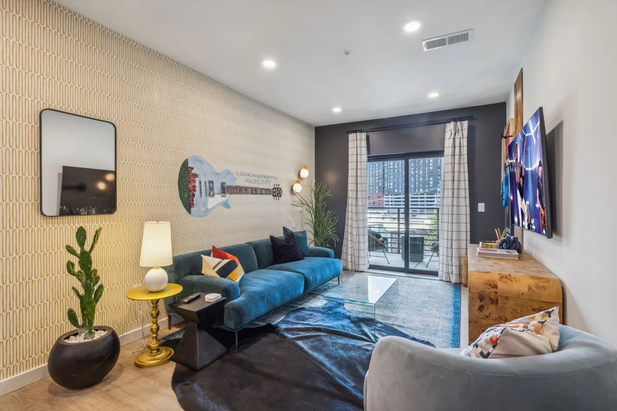 Another angle of a living area featuring a blue velvet sofa, decorative guitar mural, large window with flowing curtains, and a TV setup. The space feels airy, creative, and ideal for entertaining or unwinding, further emphasizing the property’s musical and urban ambiance.