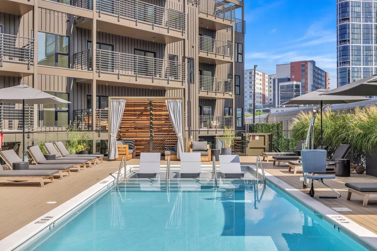 A bright exterior shot of the resort-style pool area, featuring a sparkling swimming pool, lounge chairs, umbrellas, and the building’s facade. The mood is chill and leisurely, inviting guests to unwind after a day of adventure.