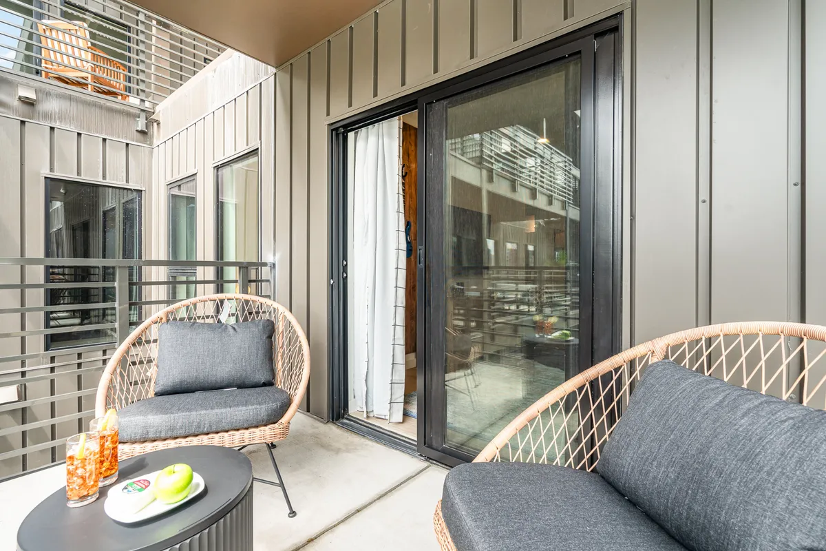 A private patio just outside the sliding glass doors, featuring wicker chairs, a small table, and a relaxed, casual setting. The patio invites guests to enjoy fresh air and intimate moments with the home as a backdrop.