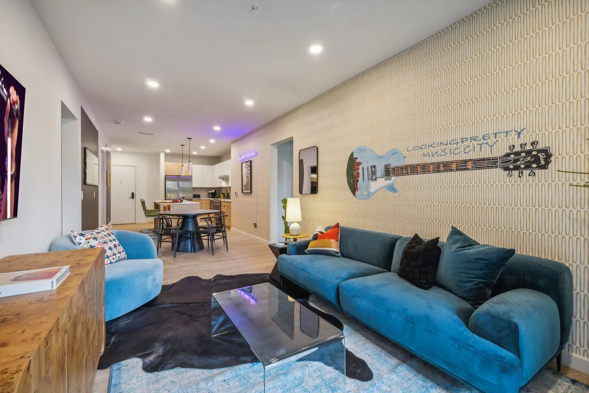 A stylish living room with a blue sofa, vibrant guitar mural, glass coffee table, and adjacent dining area. The decor is playful and artistic, evoking Nashville's music culture and the space’s creative energy. This setup encourages socializing, relaxing, or intimate music sessions, reflecting the spirit of the city.