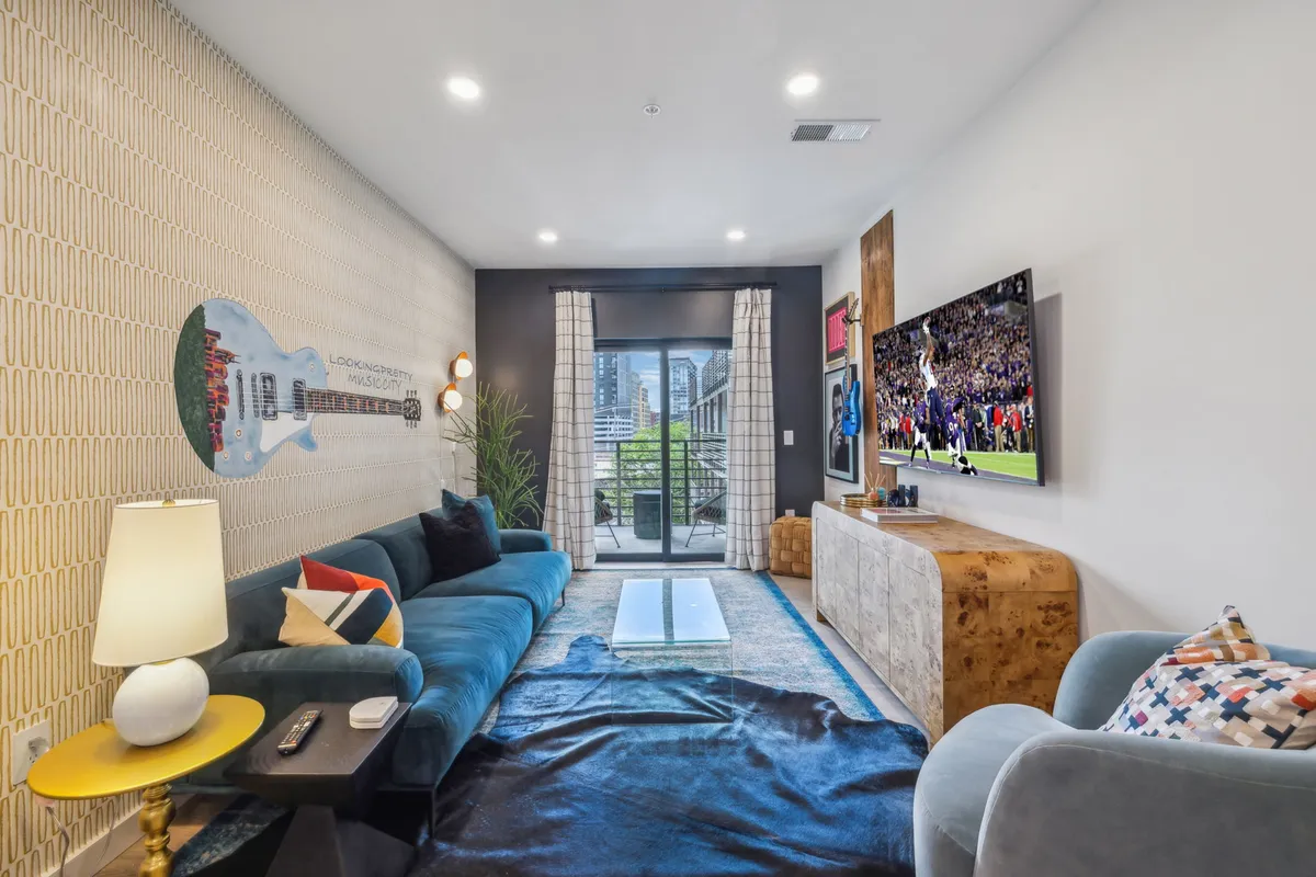 A cozy living area featuring a blue sofa, large TV, sliding glass doors leading to a balcony, and playful guitar-themed wall art. The space is bright and inviting, perfect for movie nights or relaxed gatherings with friends and family.