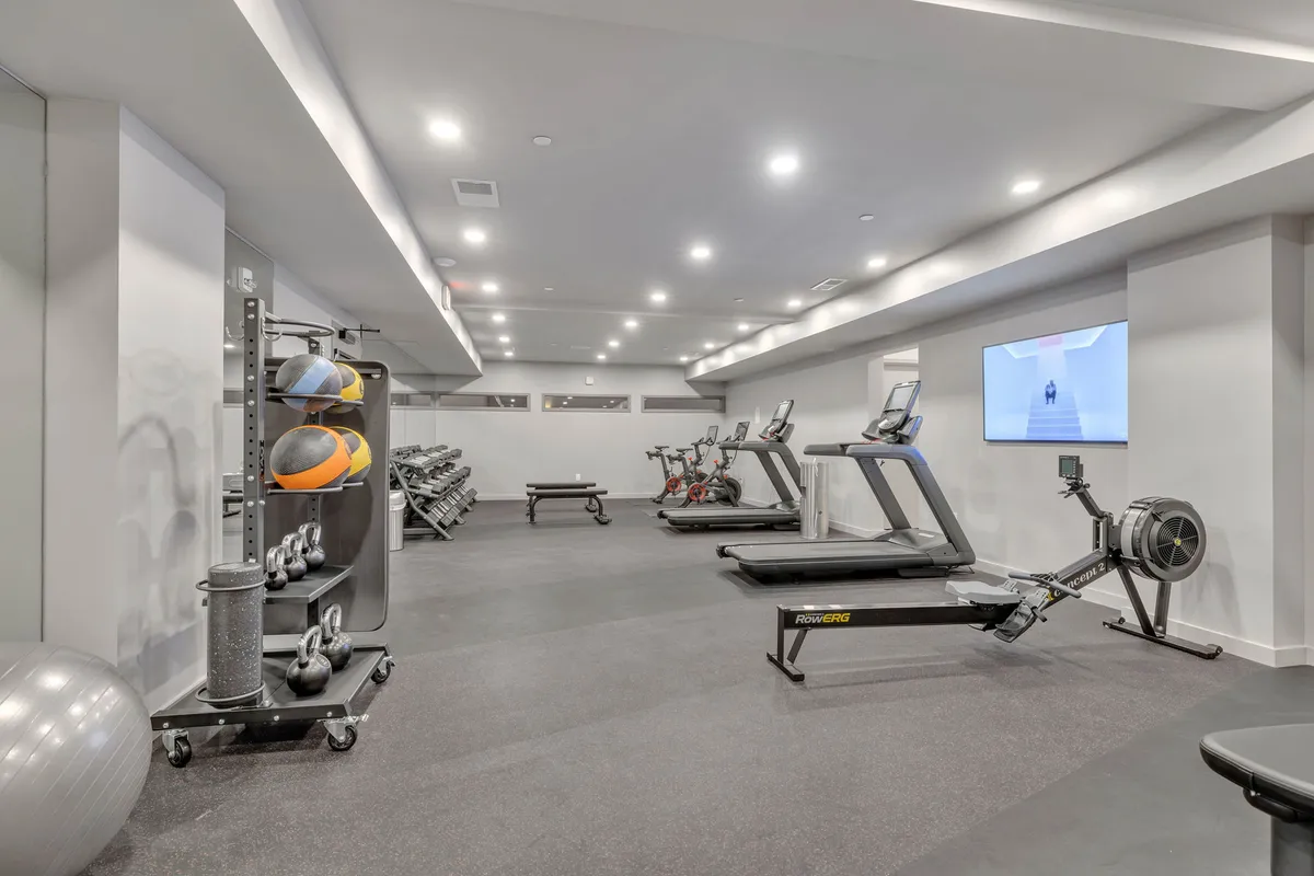 A modern, well-equipped gym interior with treadmills, exercise bikes, rowing machines, a weight bench, and medicine balls set against large windows. The space is bright, motivational, and designed for both cardio and strength training.
