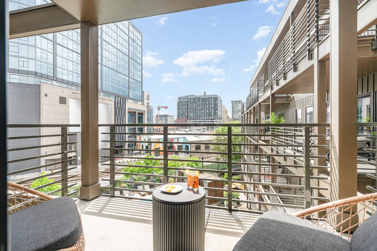 A stylish balcony seating area with a view of the cityscape and a decorative table set for drinks and snacks. The scene suggests a perfect spot for morning coffee or evening relaxation, blending indoor comfort with outdoor enjoyment.