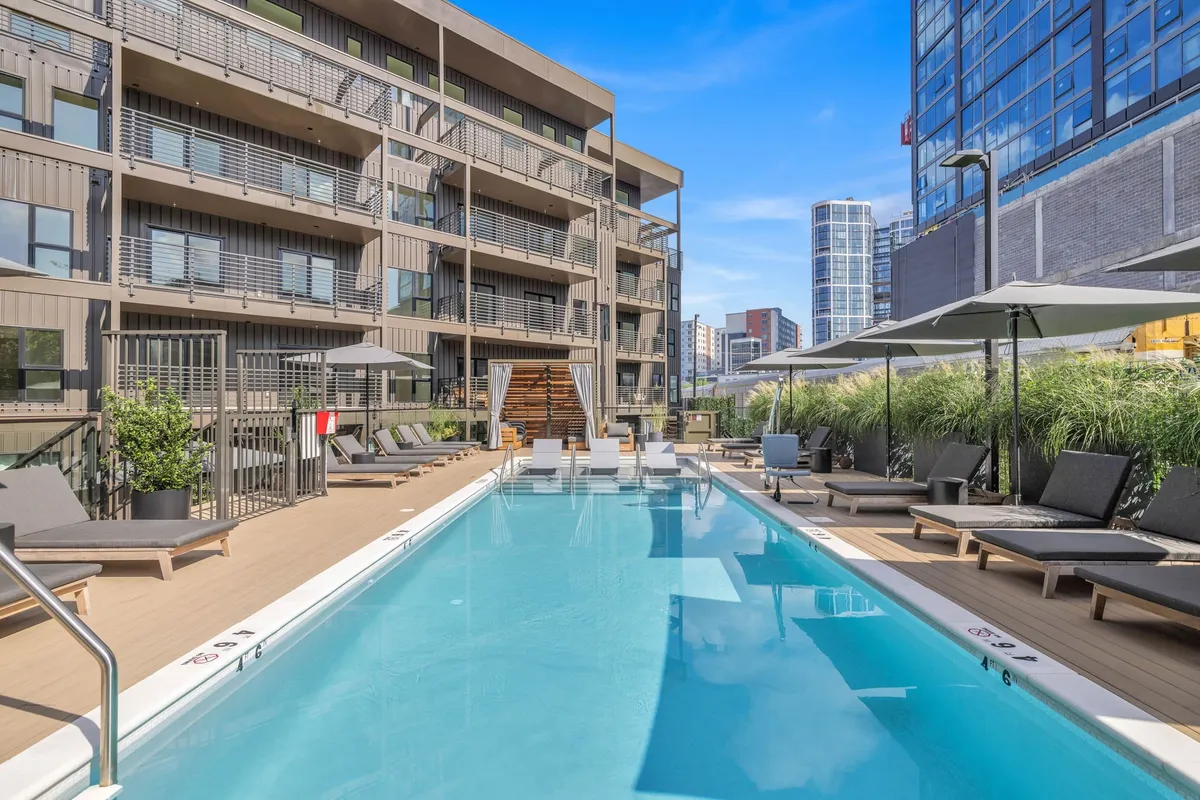 A vibrant outdoor swimming pool lined with lounge chairs and surrounded by lush greenery. The building's facade is visible in the background, and the relaxed pool area suggests both social gatherings and quiet moments. The atmosphere is lively yet tranquil, inviting guests to enjoy sunbathing, swimming, or relaxing by the water.