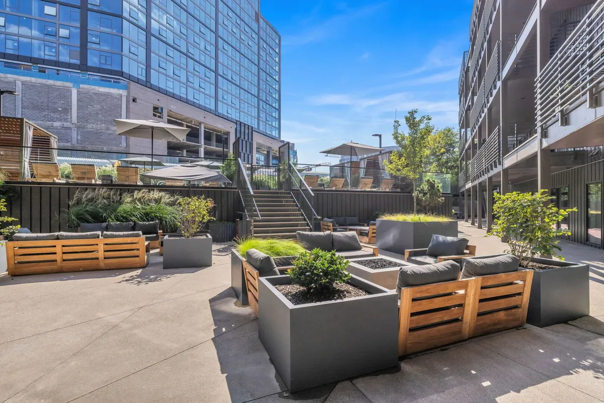 A spacious rooftop lounge featuring multiple outdoor seating areas, planters with greenery, and a staircase leading to a higher level. The atmosphere is relaxed and communal, ideal for sunset views, social events, or quiet evenings by the firepit.