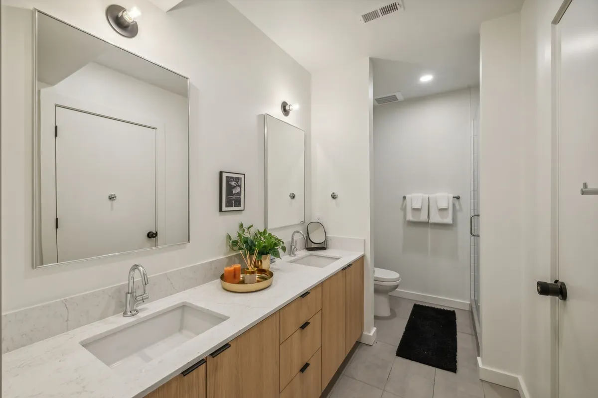 A spacious, modern bathroom with dual sinks, large mirrors, wooden cabinetry, a decorative plant, and neatly arranged towels. The space feels refreshing, perfect for self-care routines and unwinding after a long day.